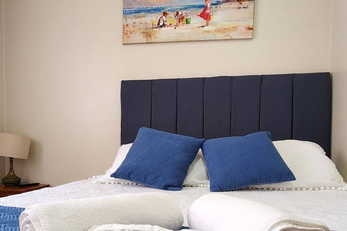 Una cama bien hecha con ropa de cama azul y blanca, un cuadro de una escena de playa en la pared y una lámpara a un lado.