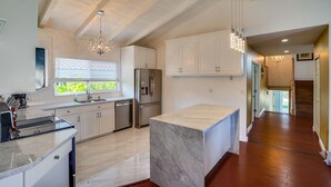 A modern kitchen with white cabinets, a marble island, stainless steel appliances, and a large window with blinds.
