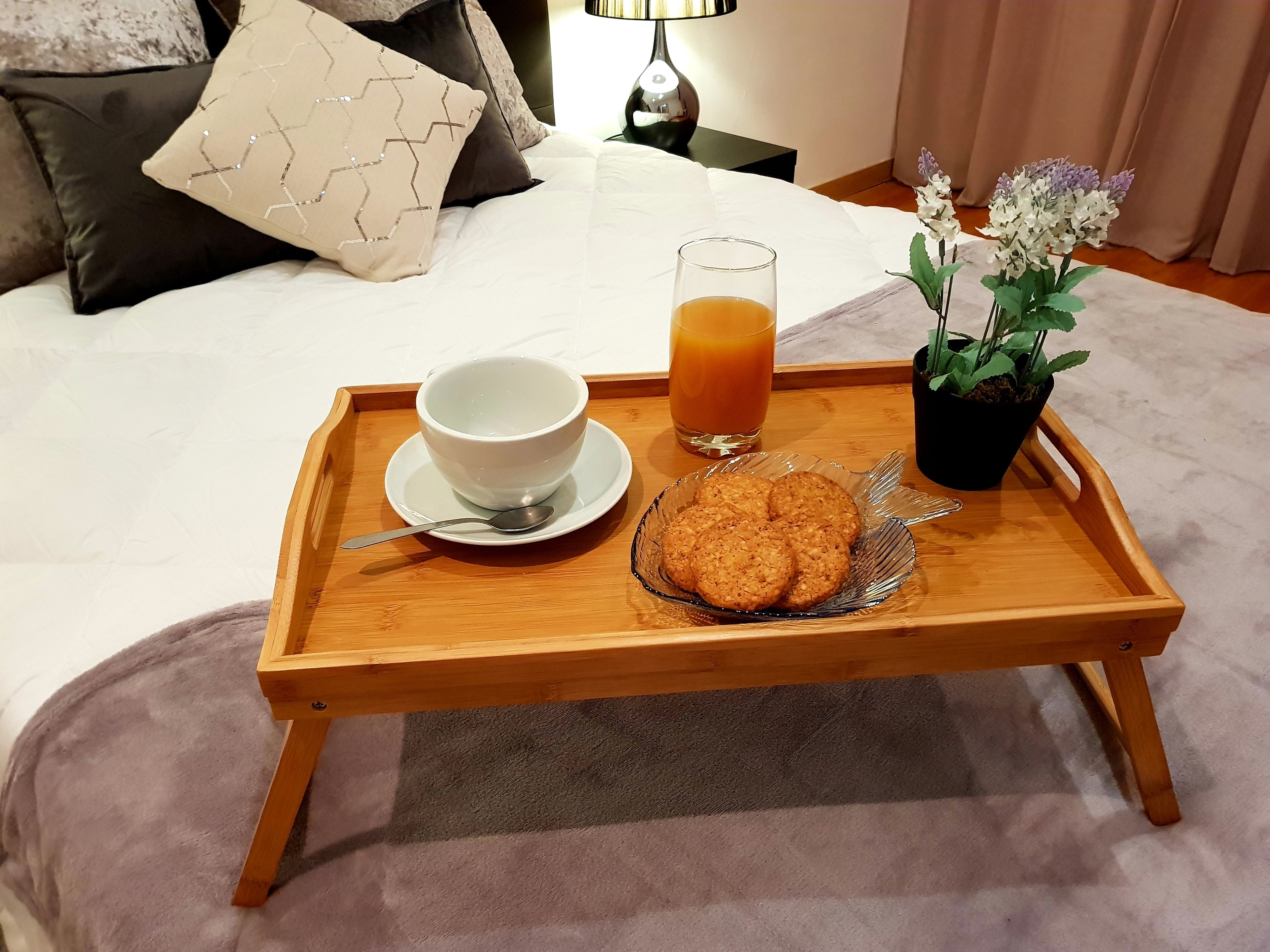 Un plateau en bois avec un bol, un verre de jus d’orange et une assiette de biscuits posés sur un lit.