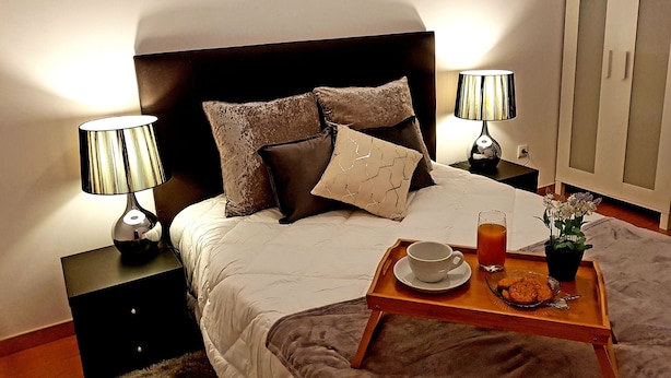 A neatly made bed with a headboard, two bedside lamps, a tray with a cup and a glass of juice, and a wardrobe in the background. - Enjoy Oporto Flat (Porto)