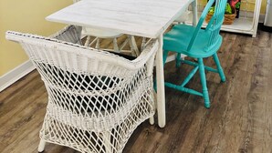 Ein Zimmer mit einem weißen Korbstuhl, einem weißen Tisch und einem Türkis-farbenen Sessel. Es gibt ein Regal mit verschiedenen Gegenständen, darunter eine rote Kaffeemaschine und ein Gemälde.