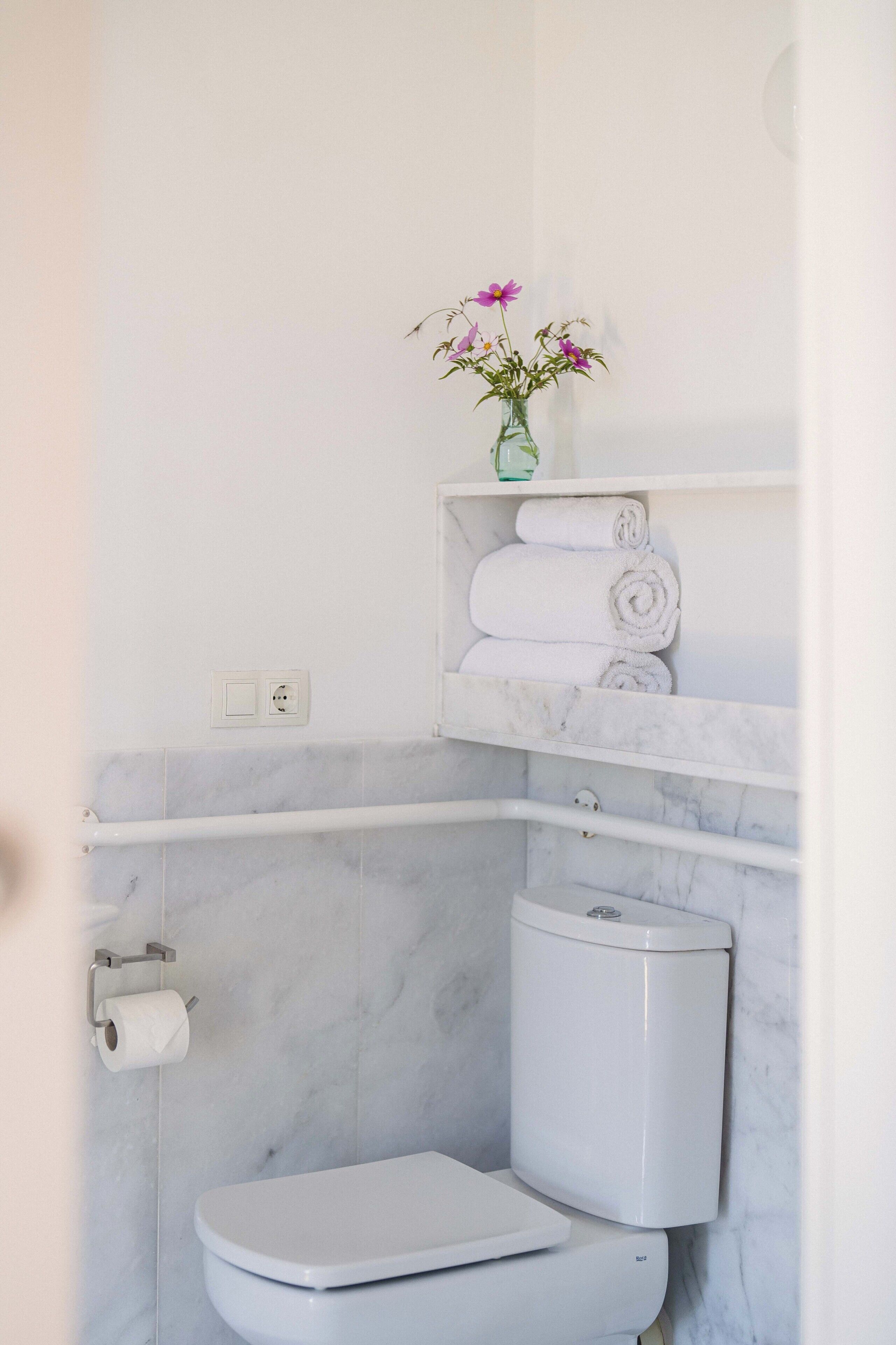 A bathroom with a toilet, marble walls, a towel rack, and a vase with flowers.