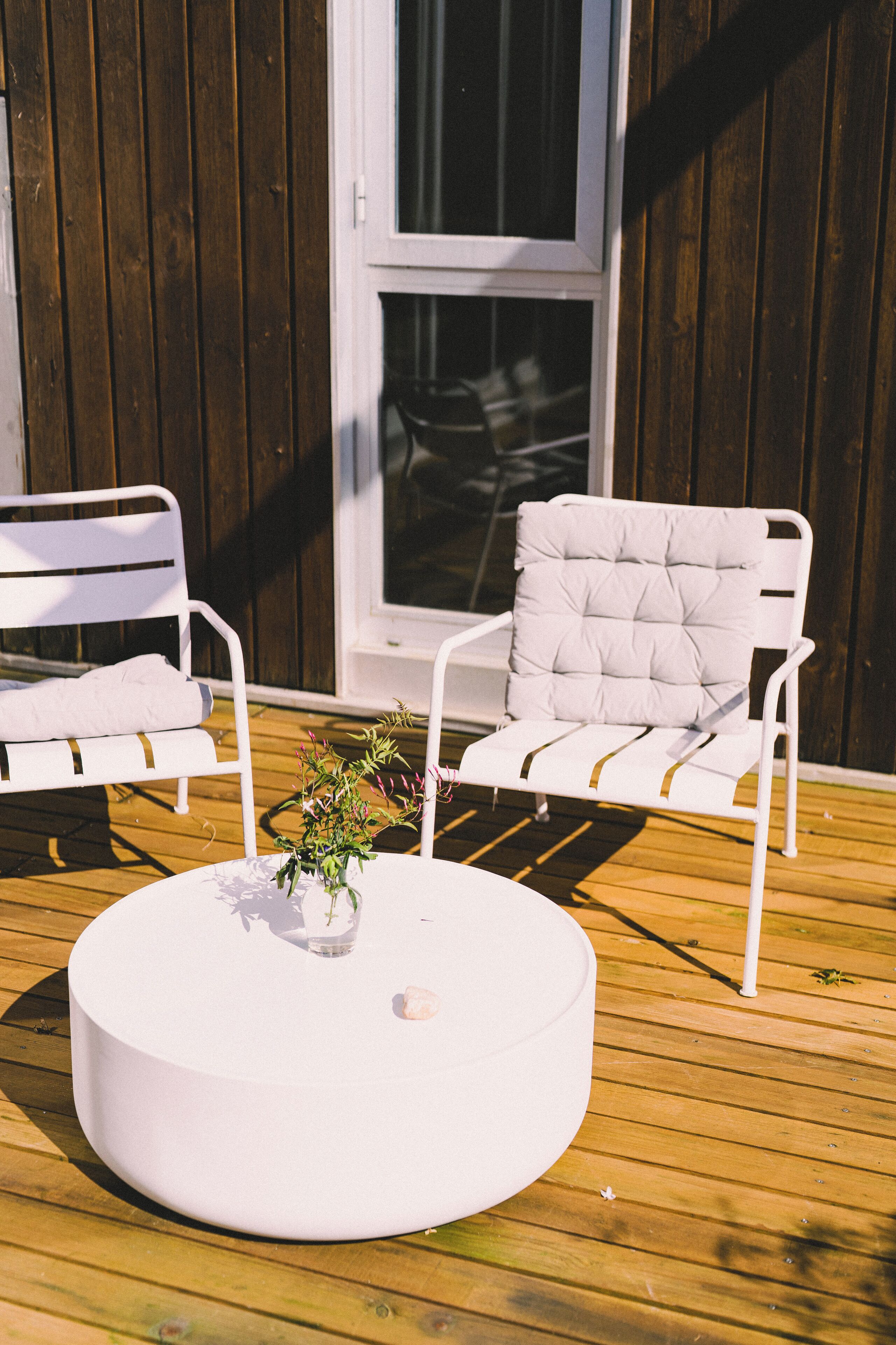 A round white table with a potted plant in the center, two white chairs with cushions, and a wooden deck.