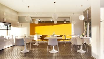 Un comedor moderno con mesas redondas y sillas blancas, una pared de acento amarilla e iluminación colgante.