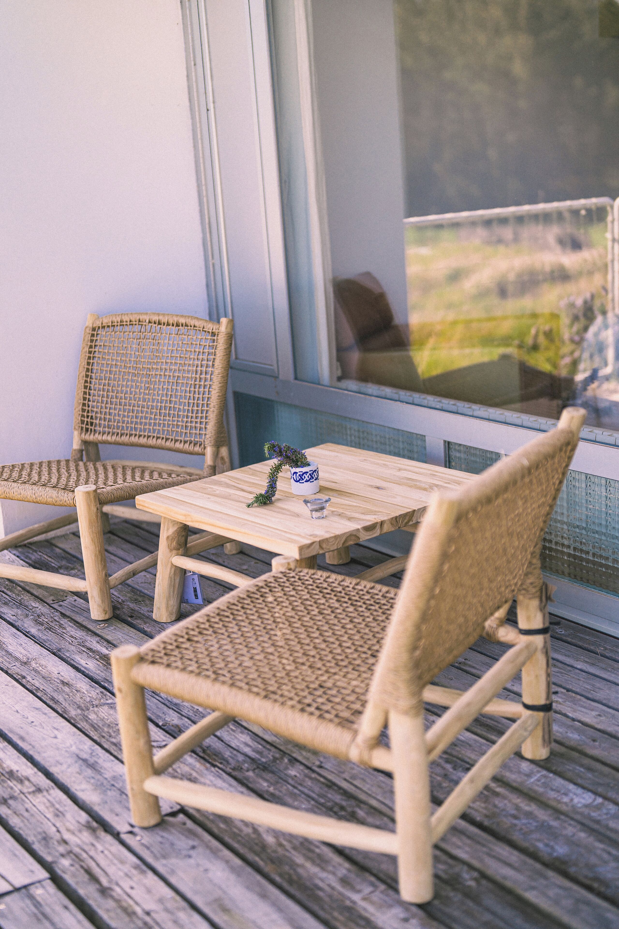 Eine Holzterrasse mit zwei Korbstühlen und einem kleinen Tisch, ein Fenster mit Blick ins Grüne und eine Glastür.