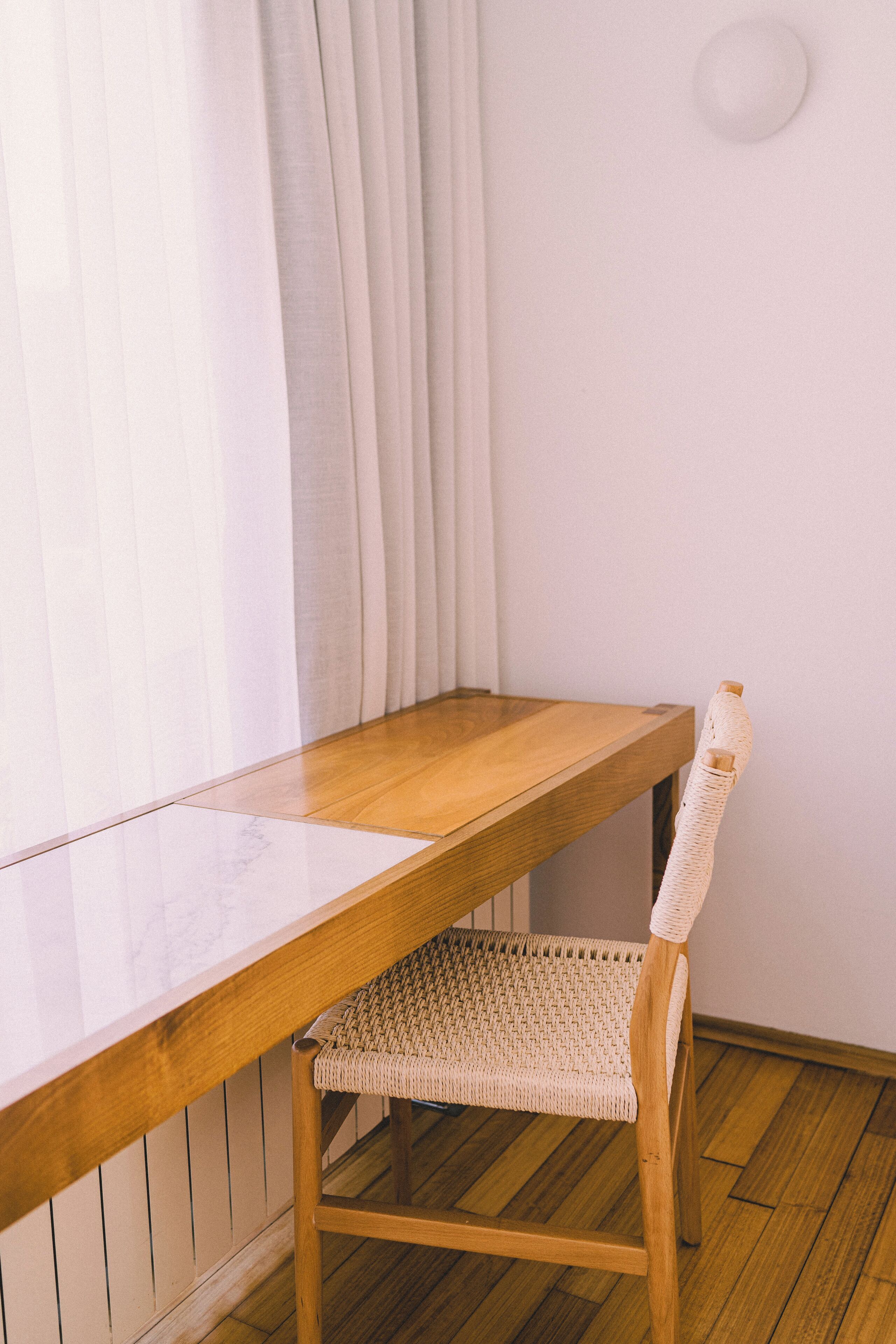 Un escritorio de madera con una silla trenzada en una habitación con suelo de madera y paredes blancas.