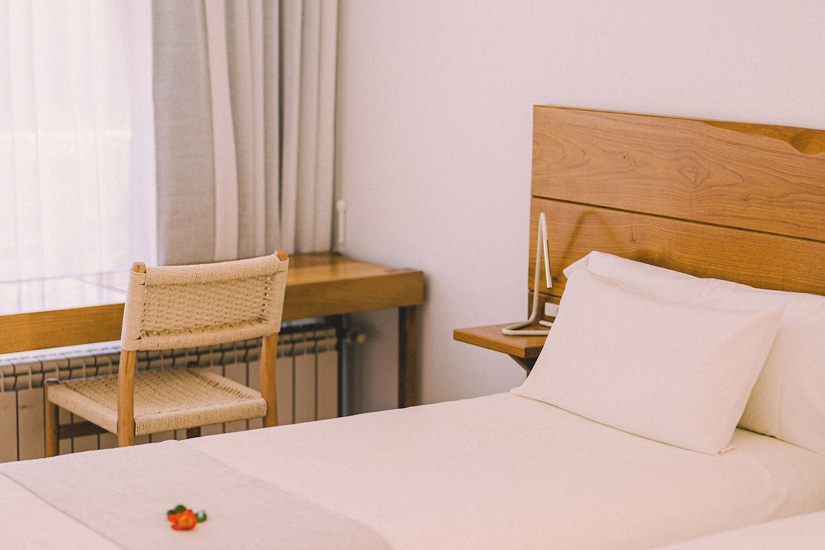 Quarto de hotel com duas camas, uma cabeceira de madeira, uma mesinha, uma cadeira e uma janela com cortinas.