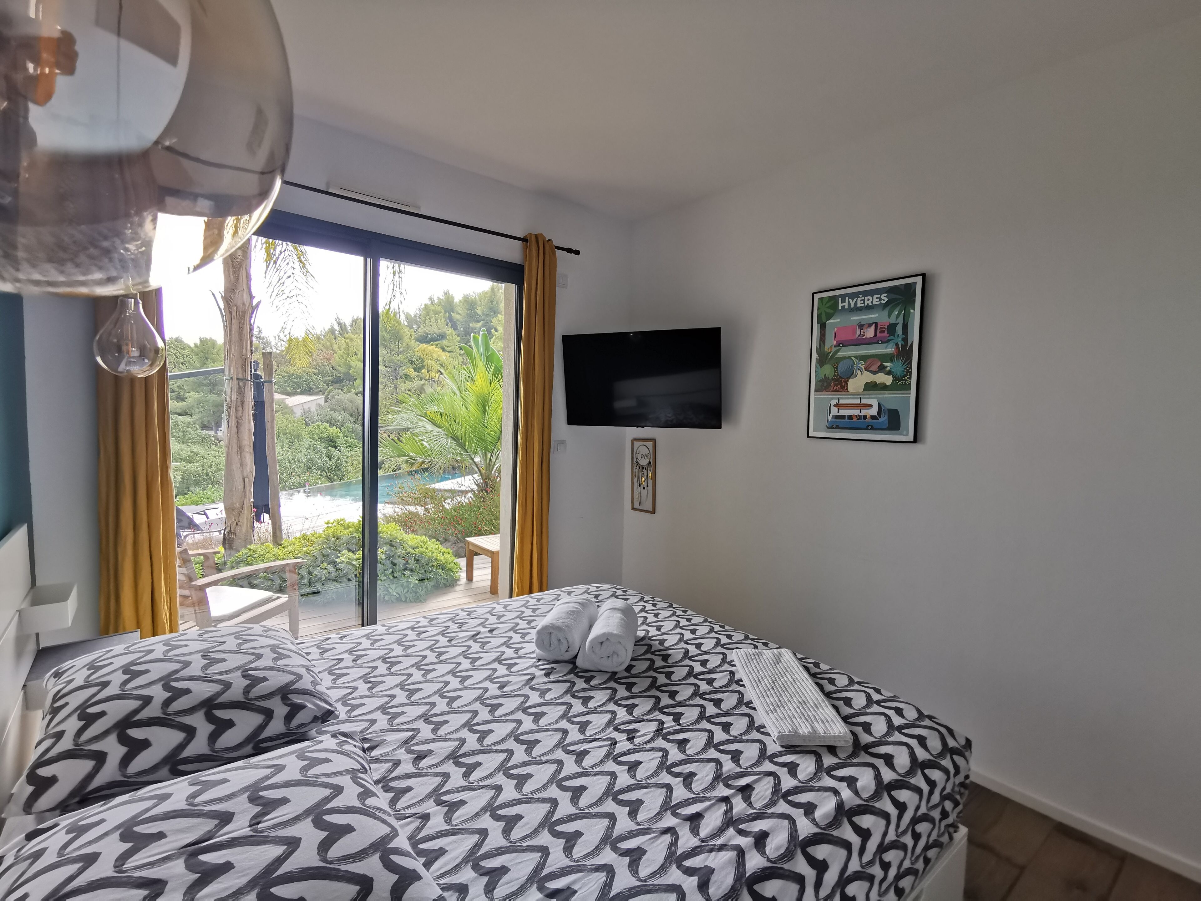 A bedroom with a patterned bedspread, a flat-screen TV, a framed picture, and a view of a swimming pool and greenery outside.
