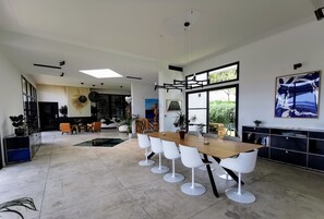 A spacious, modern dining area with a long wooden table and white chairs, surrounded by a minimalist kitchen and a large window. - Exceptional property with sea view - swimming pool - spa - gym. (Hyères)