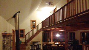 A spacious living room with a high ceiling, a wooden staircase, and a kitchen area visible through an open doorway. - Waterfront Lakehouse (Stetson)
