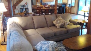 A spacious living area with a large sectional sofa, a patterned throw pillow, and a small round table. A staircase leads to an upper level, and a dining area is visible in the background. - Waterfront Lakehouse (Stetson)