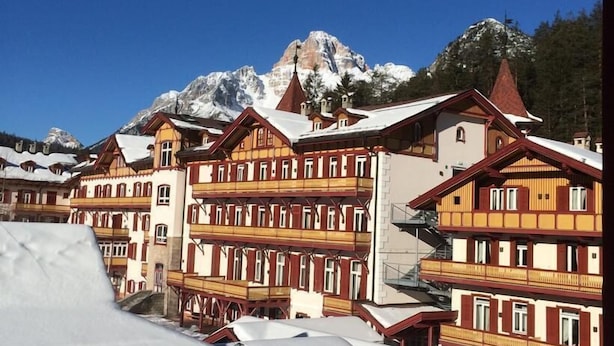 Una hilera de edificios de estilo chalete tradicional con balcones, con montañas nevadas al fondo bajo un cielo azul despejado.