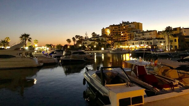 A marina at dusk with boats docked, a large building with multiple floors, and palm trees in the background. - Cabopino (Marbella)