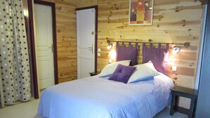 Une chambre avec un mur en bois, un lit recouvert d’une courtepointe blanche et orné d’oreillers violets, deux tables de chevet avec des lampes, et une porte avec un rideau.