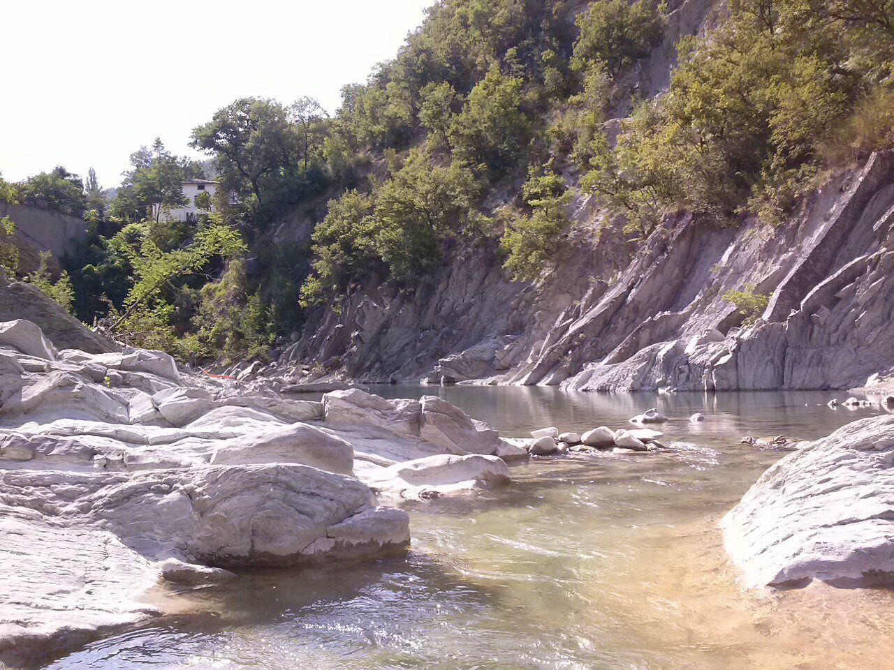 A natural rocky landscape with a river, trees, and a cliff.