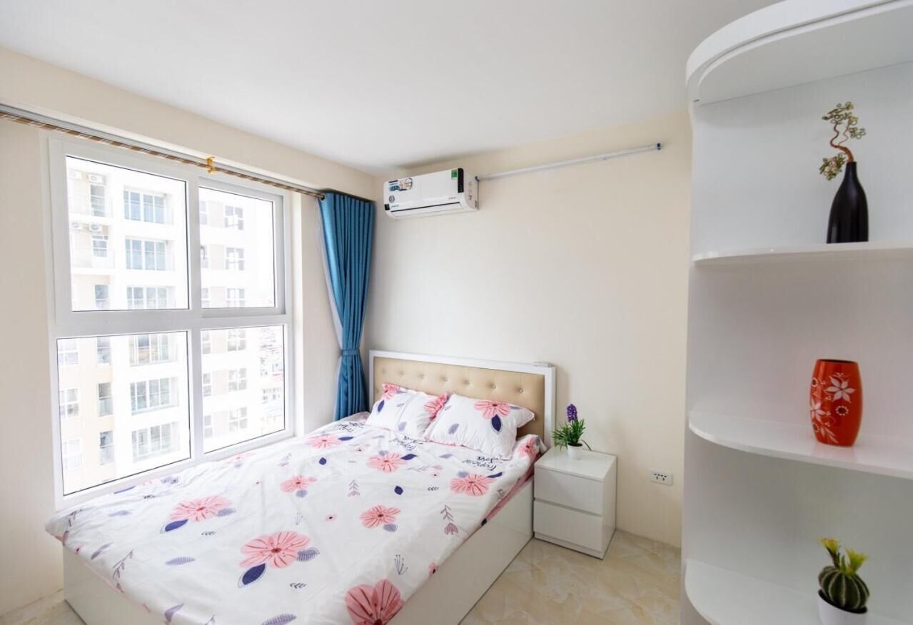 Une chambre à coucher comprenant un lit, une table de chevet, un climatiseur fixé au mur, une fenêtre avec des rideaux et une étagère décorative.
