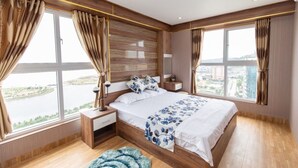 A modern bedroom with a large bed, wooden headboard, and a view of a cityscape through large windows. - Ha Long Pearl Apartment (Bai Chay)