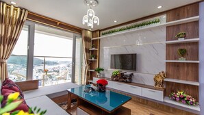 A modern living room with a large TV, wooden shelves, and a view of the city. - Ha Long Pearl Apartment (Bai Chay)
