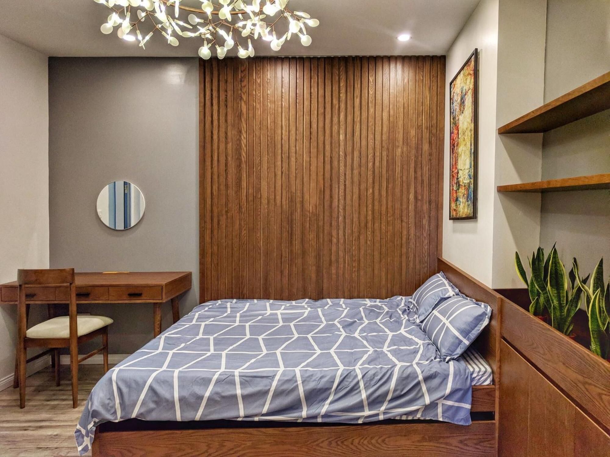 Un dormitorio con cabecera de madera, cama con colcha de estampado geométrico, espejo redondo, escritorio con silla y una planta en maceta.