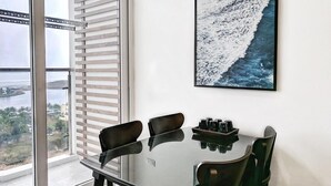 A dining area with a glass table, four chairs, and a view of a body of water through a window with blinds. - Ha Long Pearl Apartment (Bai Chay)