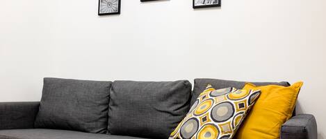 A grey sofa with two cushions, one yellow and one with a geometric pattern, against a white wall with several framed pictures.