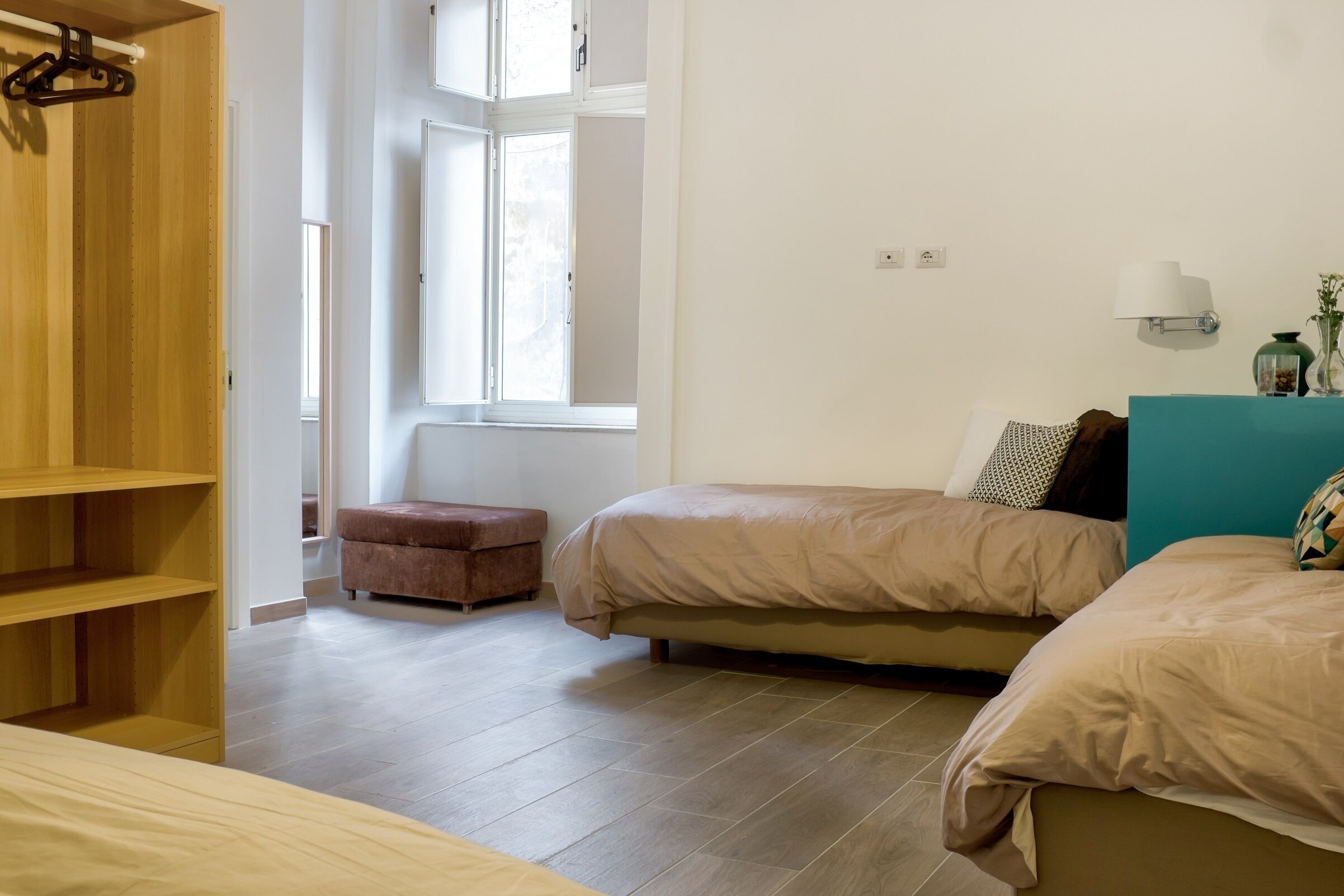 A bedroom with two beds, a wooden wardrobe, a window, and a bedside table with a lamp.