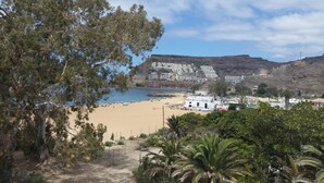 Una spiaggia con un edificio bianco con la scritta 'BARTOPIO' in primo piano, una spiaggia sabbiosa e una collina rocciosa sullo sfondo.