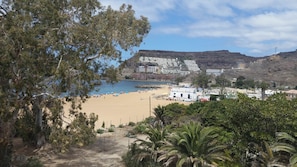 Une plage avec un bâtiment blanc sur lequel on peut lire l’inscription « BARTOPIO » au premier plan, une plage de sable et une colline rocheuse en arrière-plan.
