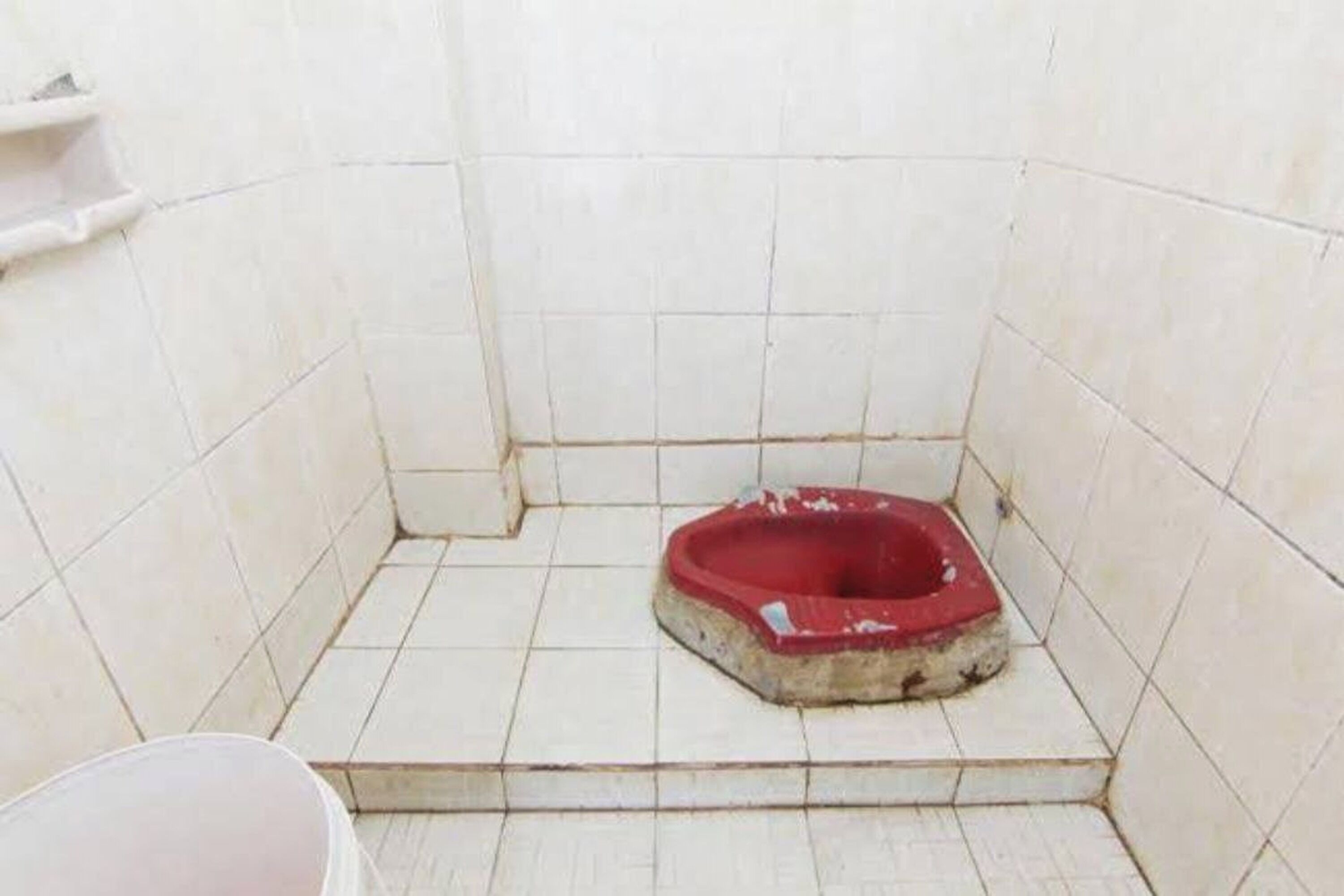 A small, tiled bathroom with a red toilet and a white bucket.