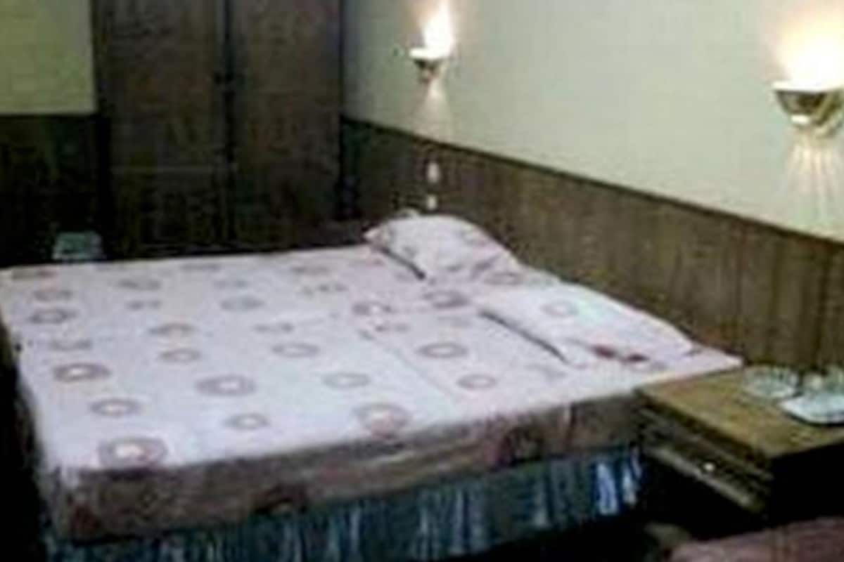 A small, single-bed room with a wooden headboard, a bedside table with a lamp, and a wall-mounted light fixture.
