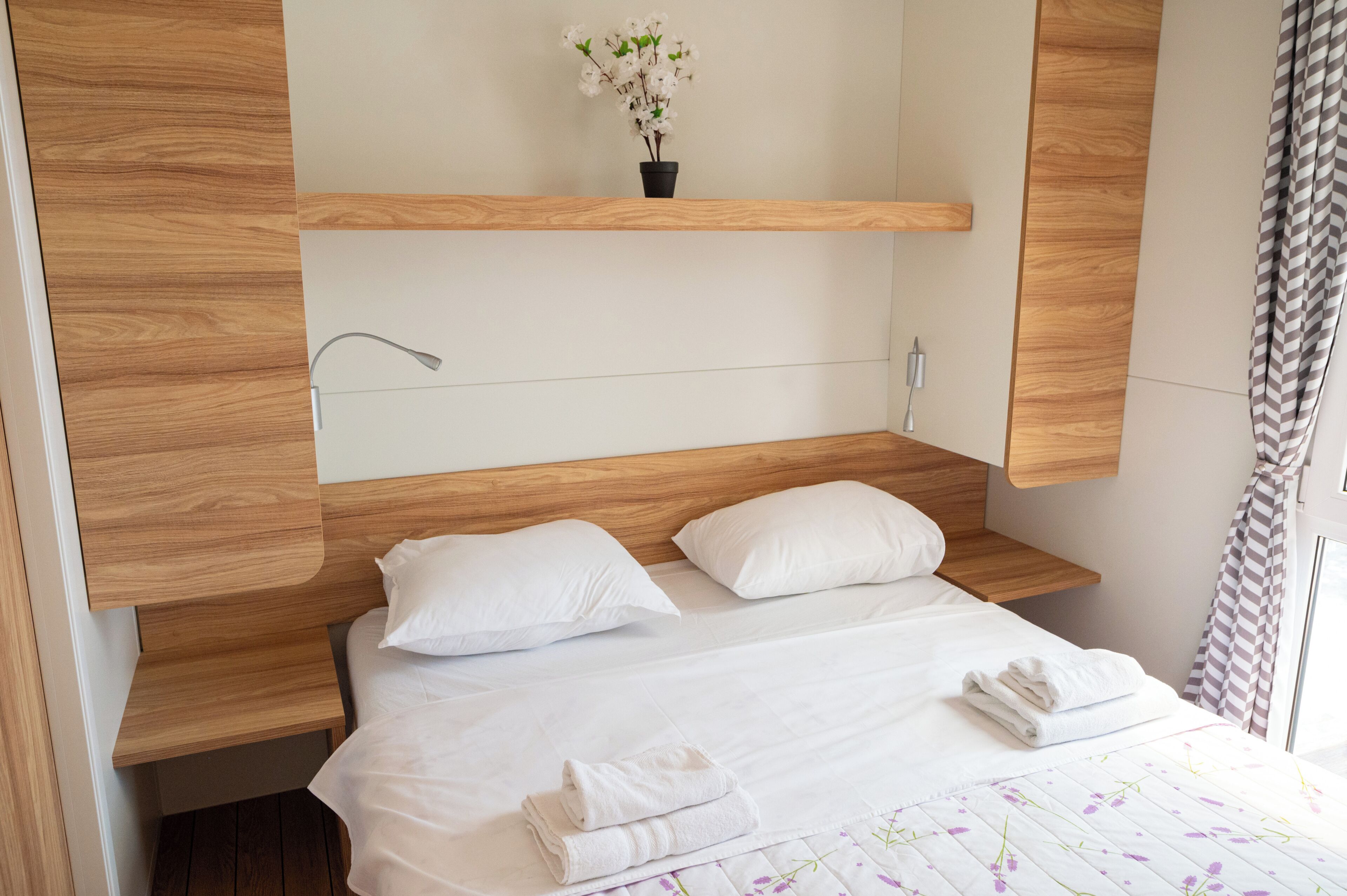 A hotel room with a wooden headboard, a bedside table, a wall-mounted lamp, a potted plant, and neatly folded towels on the bed.