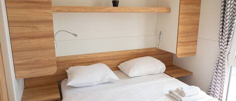 A hotel room with a wooden headboard, a bedside table, a wall-mounted lamp, a potted plant, and neatly folded towels on the bed.