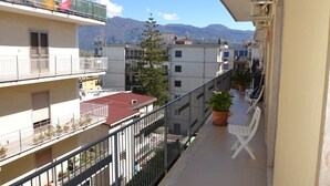 Ein Balkon mit Topfpflanzen, einem weißen Stuhl und Blick auf benachbarte Gebäude und Berge.