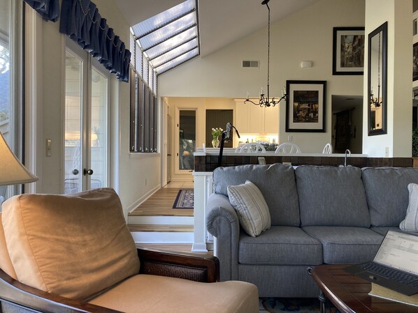 A modern living room with a grey sofa, a brown armchair, and a wooden coffee table. A laptop is on the table. The room has large windows with blinds, a chandelier, and framed artwork on the walls. - Wonderful Golf Views! Privacy, Pets, 1/2 mile to Beach, Renovated. 5Star Reviews (Hilton Head Island)