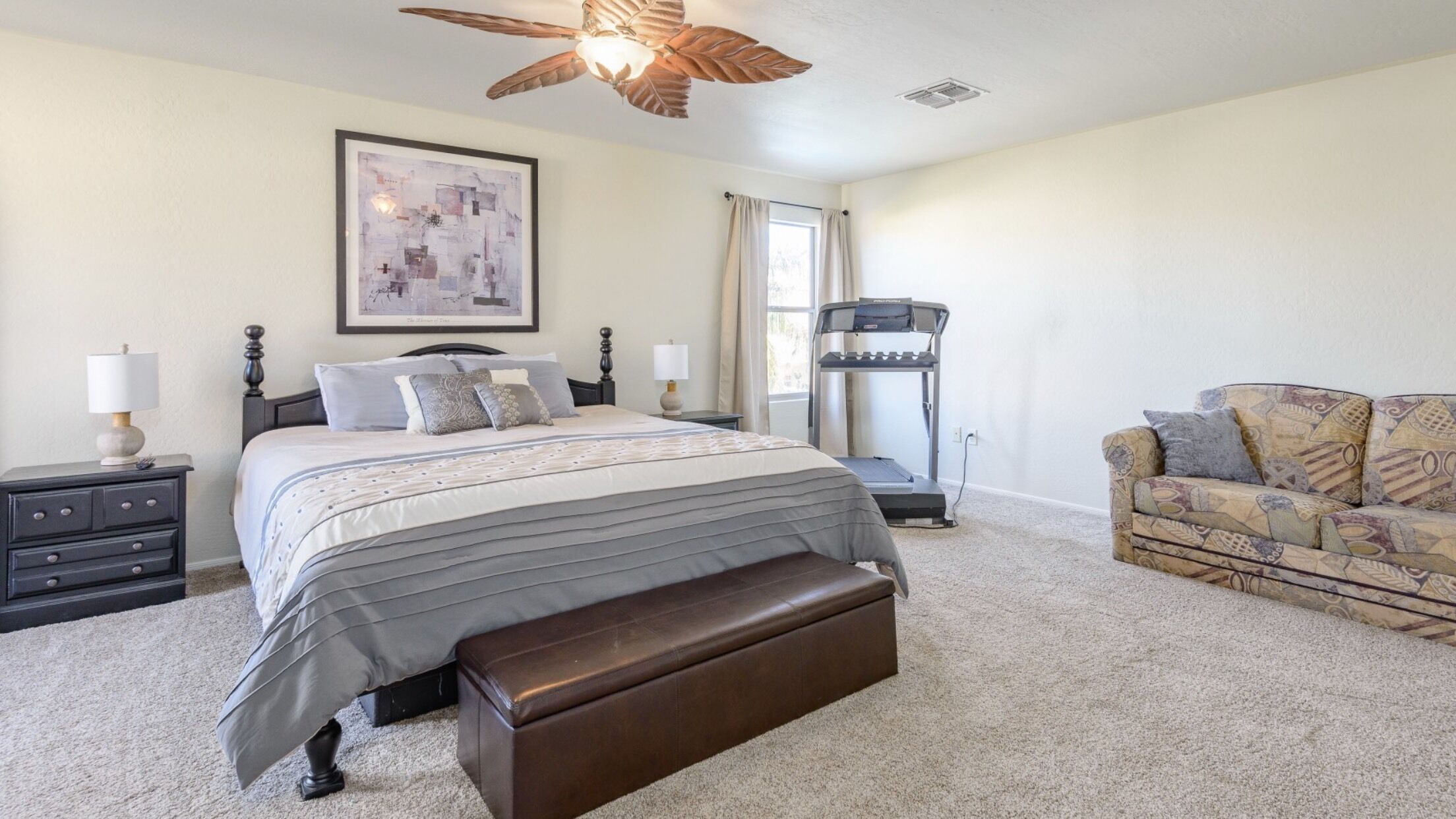A bedroom with a bed, a treadmill, a patterned sofa, and a ceiling fan.