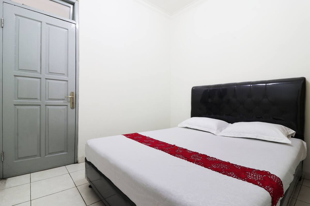A single bed with a white mattress and a red blanket, a grey door, and a white wall.