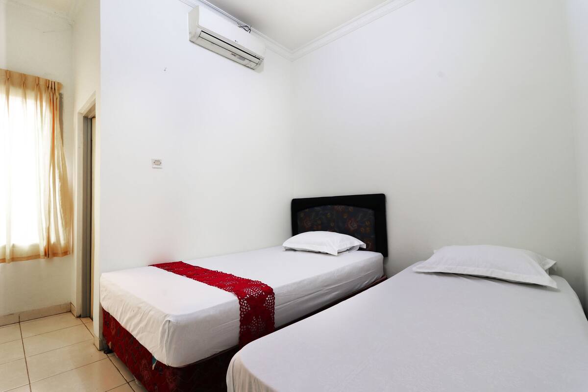 A room with two single beds, white bedding, a red decorative item on one bed, a window with curtains, and an air conditioning unit on the wall.