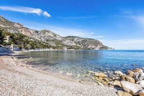 A pebble beach with clear water, rocky outcrops, and a mountainous backdrop.