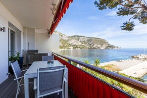 A balcony with a table and chairs, a grill, and a view of the sea and mountains.