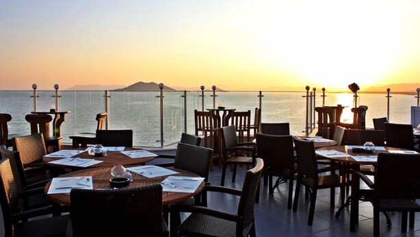 A restaurant with tables and chairs arranged for dining, overlooking a body of water and mountains in the background.