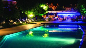 A nighttime swimming pool area with lounge chairs and a covered seating area.