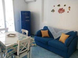 A living room with a blue sofa, two yellow cushions, a white table with a fish-patterned tablecloth, and a blue cabinet. - "AFRODITE" APARTMENT ONLY 250 MT FROM THE SEA (San Vito Lo Capo)