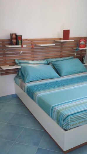A bed with a striped blue and white bedspread, two pillows, and a wooden headboard with shelves holding books and a red object. - "AFRODITE" APARTMENT ONLY 250 MT FROM THE SEA (San Vito Lo Capo)