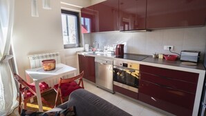 A modern kitchen with red cabinets, a dishwasher, an oven, and a dining area with a table and chairs. - Desire City Center Apartment Terazije (Belgrade)