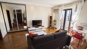 A modern living room with a large flat-screen TV, a sofa, a dining table with chairs, and a sliding glass door leading to a balcony. - Desire City Center Apartment Terazije (Belgrade)