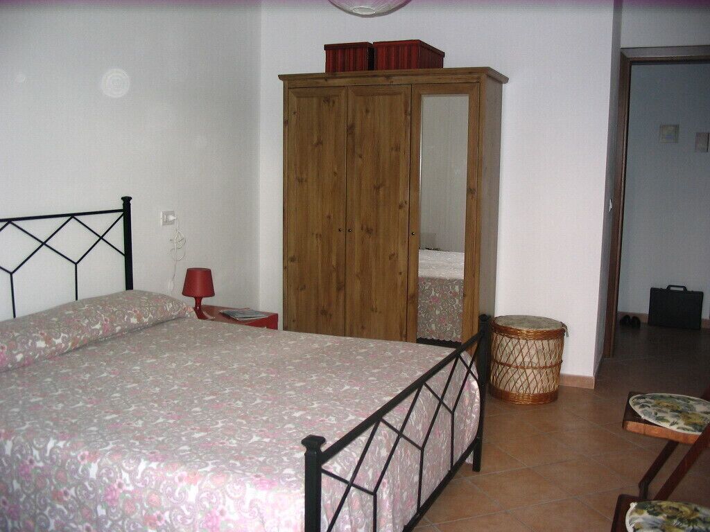 Een slaapkamer met een bed, een kledingkast, een nachtkastje en een klein rond tafeltje met een stoel.
