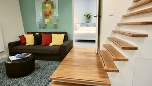 Et moderne hotelværelse med et træbord, en sofa med farverige puder, mosaikvægkunst, en trappe med trætrapper og en seng med en potteplante.