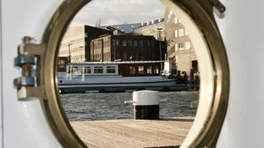 Udsigt gennem et skibs skydespil af en træbrygge, en hvid båd og bygninger langs vandet.