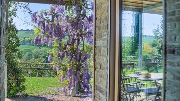 A view through a glass door of a patio area with a table and chairs, a pergola with purple flowers, and a stone wall. - Fully private fenced villa with pool – ideal for families, pet friendly (Ostra)