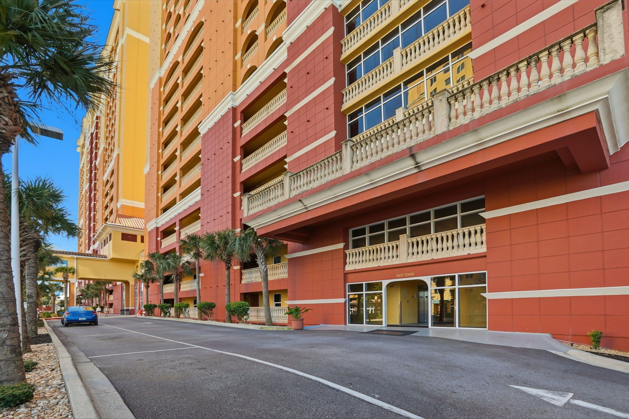 Un hotel de varios pisos con balcones, una zona de estacionamiento y palmeras.